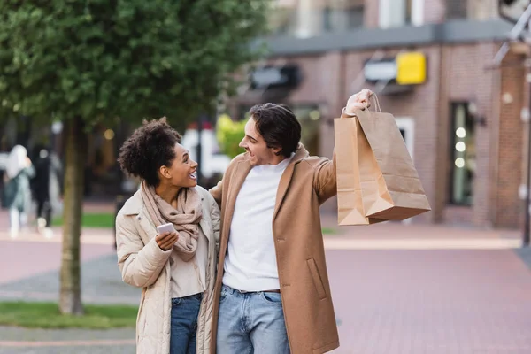 Изумленная африканская американка со смартфоном смотрит на счастливого парня, держащего сумки с покупками — стоковое фото