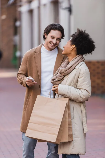 Felice uomo smartphone sorridente vicino felice donna africana americana in possesso di borse della spesa — Foto stock