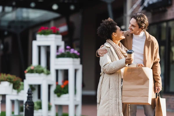 Feliz pareja multiétnica sosteniendo taza de papel y bolsas de compras mientras se miran entre sí - foto de stock