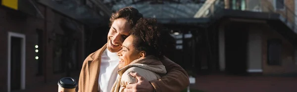 Uomo allegro che abbraccia felice donna africana americana con tazza di carta nel centro commerciale, banner — Foto stock