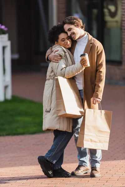 Full length of cheerful man hugging happy african american woman with paper cup and shopping bag — Stock Photo