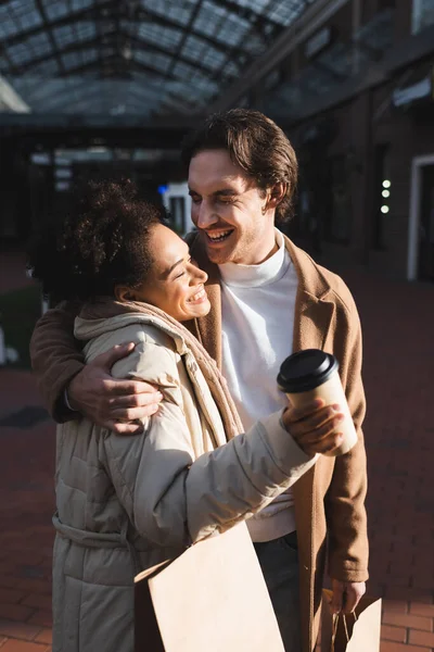 Homme souriant étreignant femme afro-américaine heureuse avec tasse en papier et sac à provisions — Photo de stock