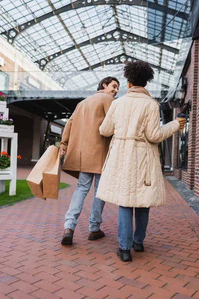 Full length of african american woman with paper cup walking with happy boyfriend holding shopping bags — Stock Photo