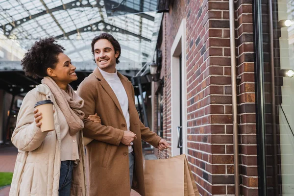 Fröhliche afrikanisch-amerikanische Frau mit Pappbecher steht mit Freund und hält Einkaufstüten am Fenster in Einkaufszentrum — Stockfoto