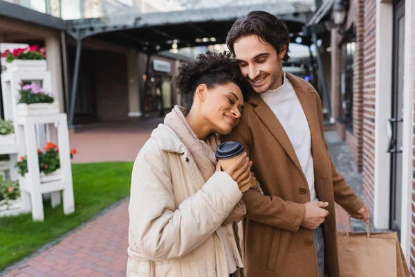 Mujer afroamericana feliz con café para ir apoyándose en novio sosteniendo bolsas de compras - foto de stock