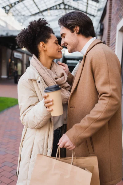 Vue latérale de femme afro-américaine heureuse avec du café pour aller près copain heureux tenant des sacs à provisions — Stock Photo