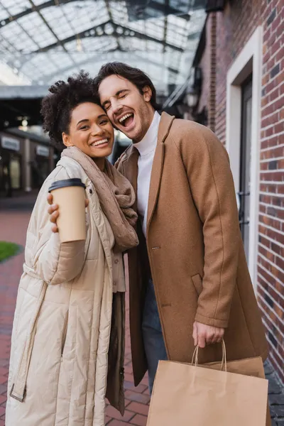 Happy african american woman holding  paper cup with coffee to go near excited boyfriend with purchases — Stock Photo