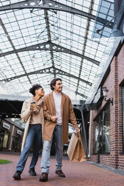 Full length of brunette african american woman with coffee to go walking near smiling boyfriend holding shopping bags — Stock Photo