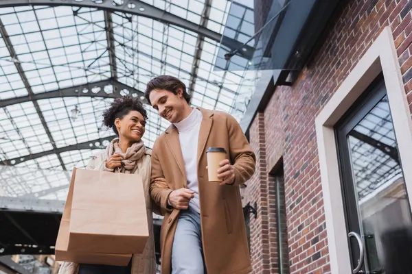 Baixo ângulo vista de feliz interracial casal segurando sacos de compras e copo de papel no shopping — Fotografia de Stock