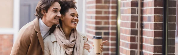 Glücklich afrikanisch-amerikanische Frau mit Pappbecher zeigt auf Gebäude in der Nähe Freund, Banner — Stockfoto