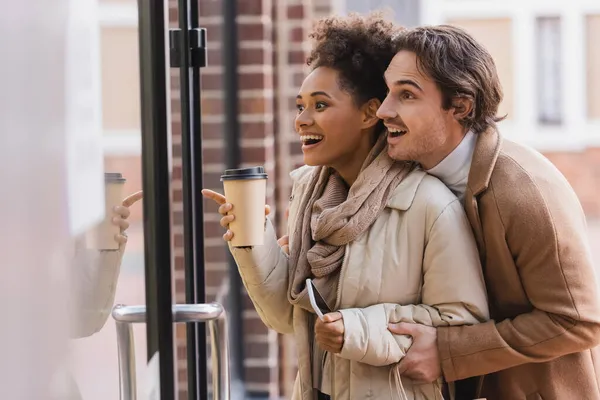 Amazed african american woman with paper cup pointing at window near boyfriend in mall — Stock Photo