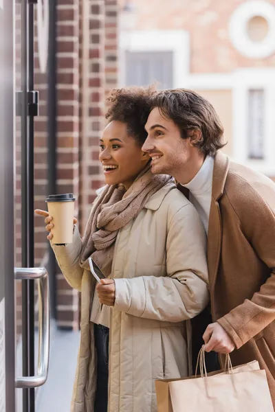Amazed african american woman with paper cup pointing at window near happy boyfriend in mall — Stock Photo