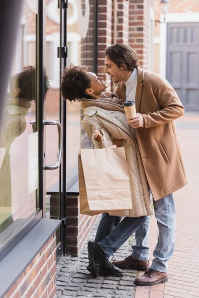 Full length of african american woman holding shopping bags and looking at happy boyfriend in coat with paper cup — Stock Photo