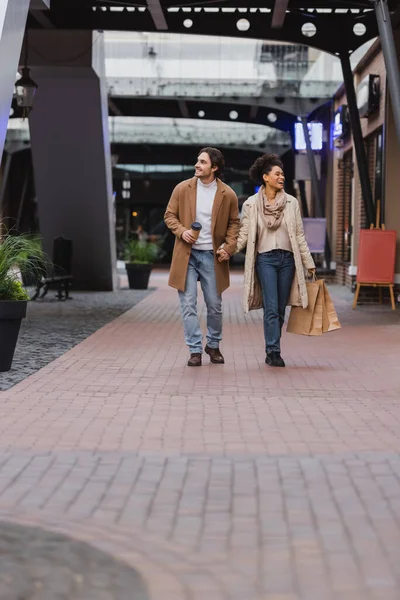 Longitud completa de la mujer afroamericana sosteniendo bolsas de compras y caminando con novio feliz en abrigo en el centro comercial - foto de stock