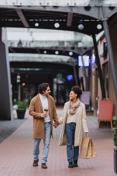 Piena lunghezza di donna afro-americana che tiene borse della spesa e cammina con gioioso fidanzato in cappotto nel centro commerciale — Foto stock