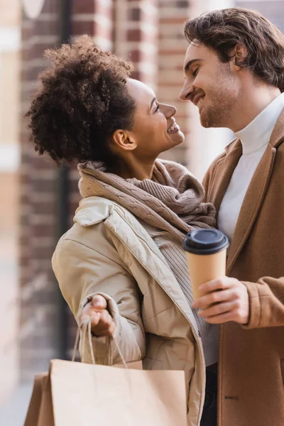 Brunette african american woman holding shopping bags and looking at happy boyfriend in coat with paper cup — Stock Photo