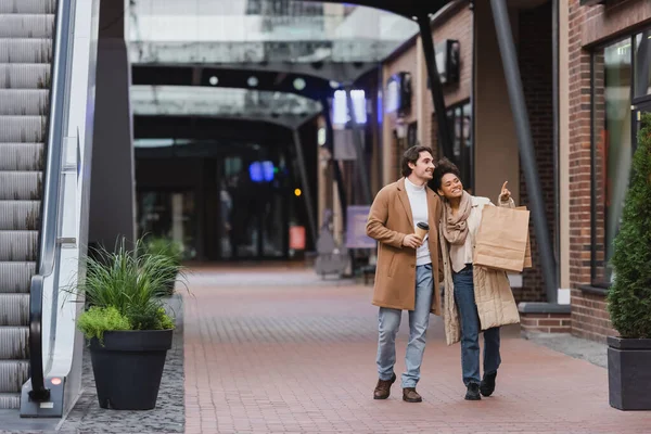 Longitud completa de la mujer afroamericana sosteniendo bolsas de compras y señalando mientras camina con novio feliz en abrigo en el centro comercial - foto de stock