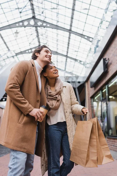 Clientes interracial sosteniendo bolsas de compras y taza de papel mientras mira hacia otro lado en el centro comercial - foto de stock