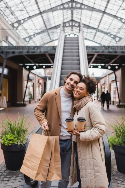 Felice donna afroamericana che tiene il caffè per avvicinarsi all'uomo con le borse della spesa sorridenti nel centro commerciale — Foto stock