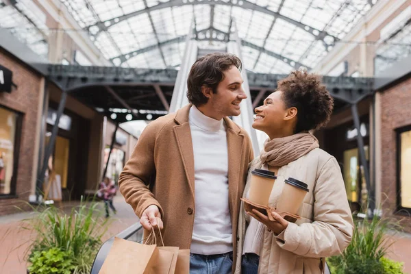 Happy african american woman holding paper cups and looking at man with shopping bags smiling in mall — Stock Photo