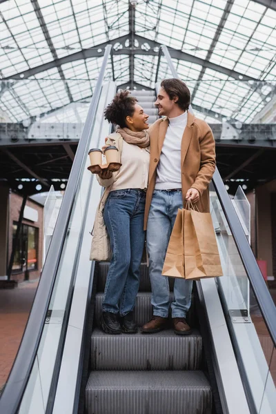 Mujer afroamericana feliz sosteniendo vasos de papel y mirando al hombre con bolsas de compras en la escalera mecánica en el centro comercial - foto de stock