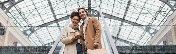 Niedrigwinkel-Ansicht der glücklichen afrikanisch-amerikanischen Frau, die Kaffee in der Hand hält, um in die Nähe ihres Freundes zu gehen, mit Einkaufstaschen auf Rolltreppe in Einkaufszentrum, Banner — Stockfoto