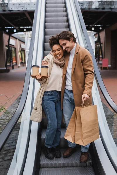Счастливая многонациональная пара с бумажными стаканчиками и сумками, обнимающая эскалатор в торговом центре — стоковое фото