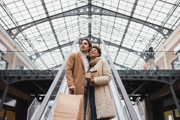 Vista de ángulo bajo de feliz pareja multiétnica con vasos de papel y bolsas de compras en escaleras mecánicas en el centro comercial - foto de stock