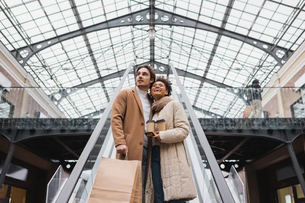 Low angle view of happy multiethnic couple with paper cups and shopping bags on escalator in mall — Stock Photo