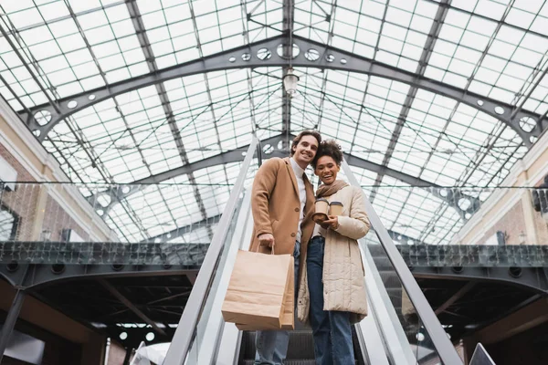 Vista de ángulo bajo de alegre pareja multiétnica con vasos de papel y bolsas de compras en escaleras mecánicas en el centro comercial - foto de stock