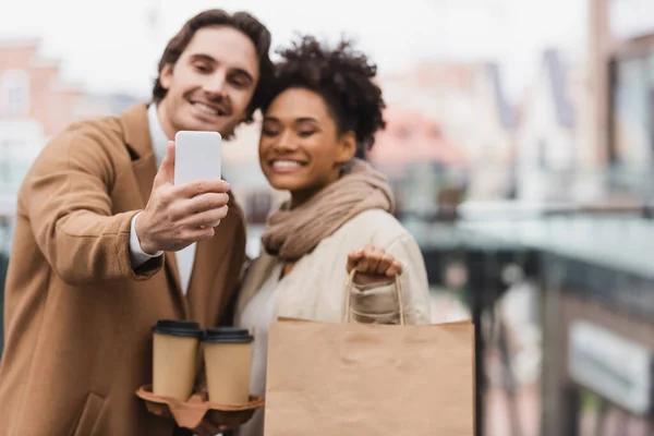 Casal multiétnico desfocado com copos de papel e saco de compras levando selfie no shopping — Fotografia de Stock