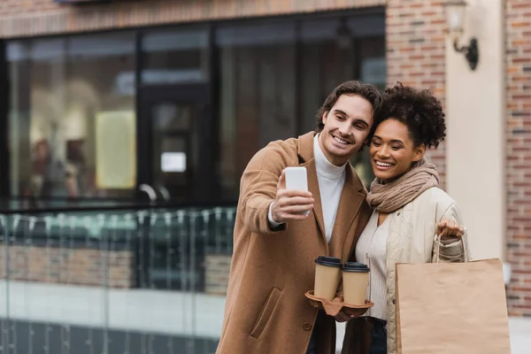 Alegre pareja multiétnica con vasos de papel y bolsas de compras tomando selfie en el centro comercial - foto de stock