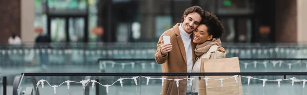 Alegre casal multiétnico com copos de papel e sacos de compras tomando selfie no shopping, banner — Fotografia de Stock