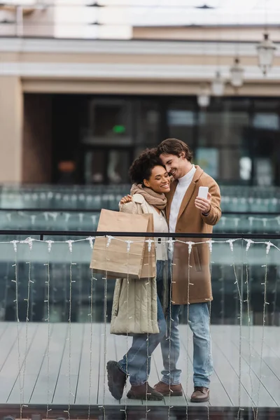 Feliz pareja multiétnica con vasos de papel y bolsas de compras tomando selfie en el centro comercial - foto de stock