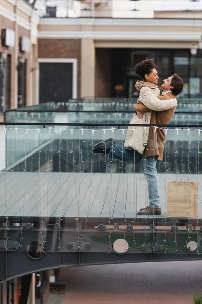 Vue de côté de l'homme heureux soulevant petite amie afro-américaine dans le centre commercial — Photo de stock