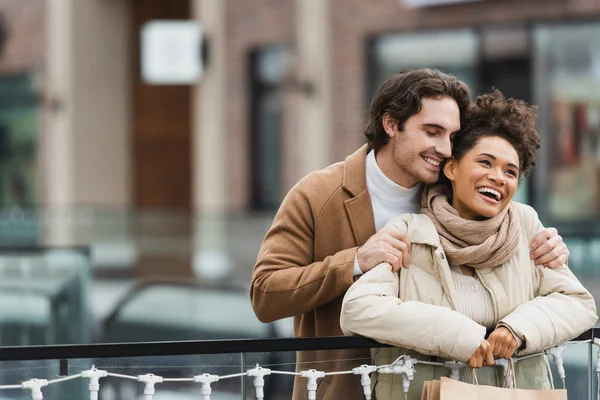 Hombre feliz abrazando a la novia afroamericana en el centro comercial - foto de stock