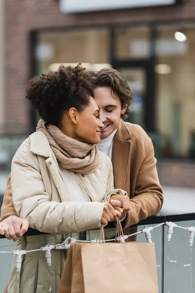 Hombre feliz cogido de la mano con la novia afroamericana sonriente en el centro comercial - foto de stock