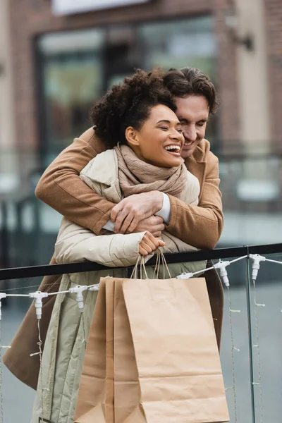 Feliz hombre abrazando alegre africana americana novia en centro comercial - foto de stock