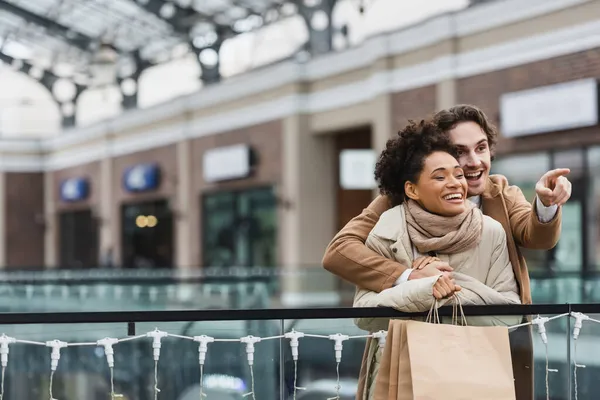 Felice uomo che abbraccia allegra fidanzata afro-americana e punta con il dito nel centro commerciale — Foto stock