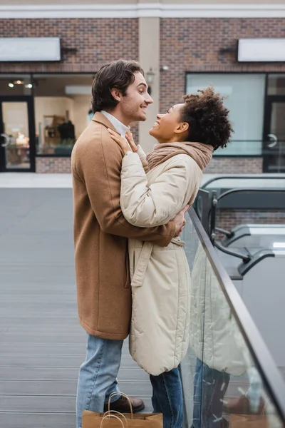 Vue de côté de l'homme heureux étreignant petite amie afro-américaine gaie dans le centre commercial — Photo de stock