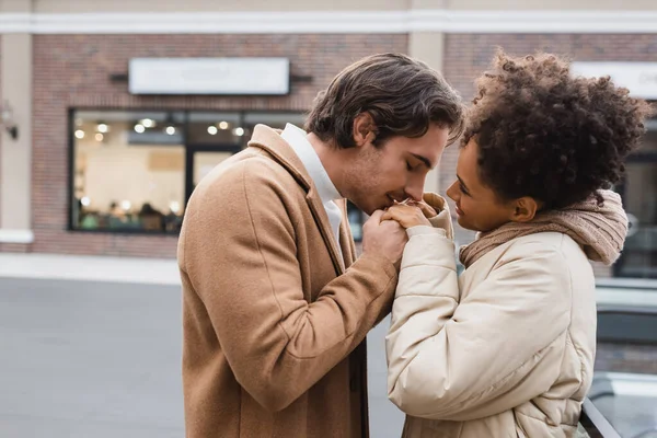 Vue de côté de l'homme heureux baisant les mains de joyeuse petite amie afro-américaine dans le centre commercial — Photo de stock