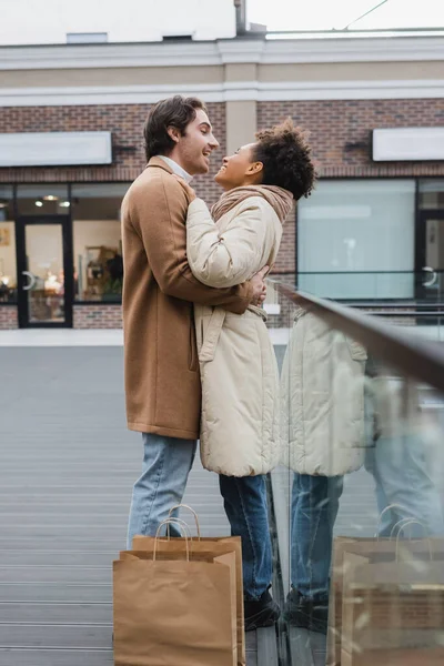 Vista lateral del hombre feliz abrazando alegre novia afroamericana cerca de bolsas de compras en el centro comercial - foto de stock