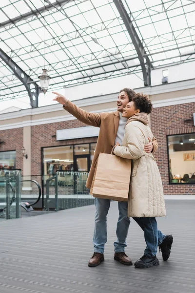 Homem feliz abraçando alegre afro-americana namorada com sacos de compras ao apontar com o dedo no shopping — Fotografia de Stock