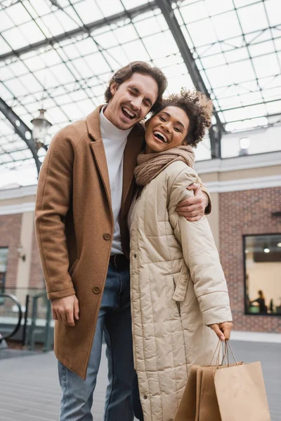 Веселый мужчина обнимает веселую африканскую американскую подружку с сумками в торговом центре — стоковое фото