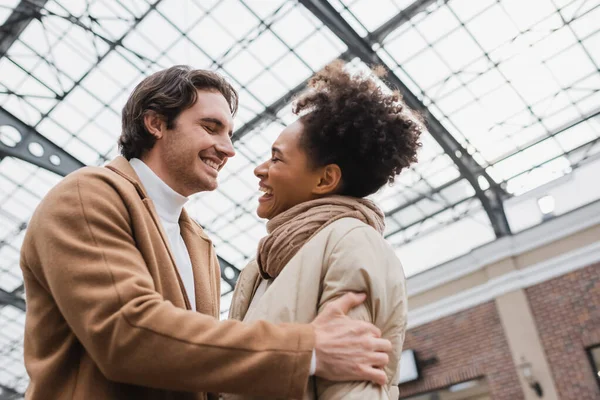 Niedrigwinkel-Ansicht des freudigen Mannes umarmt fröhliche afrikanisch-amerikanische Freundin in Mall — Stockfoto