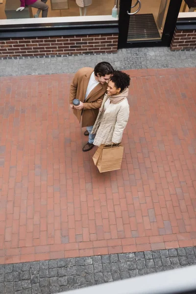 Vista superior del hombre alegre sosteniendo taza de papel cerca de la novia afroamericana complacida con compras en el centro comercial - foto de stock