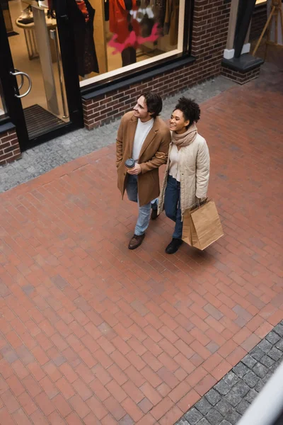 Vista de ángulo alto del hombre alegre sosteniendo taza de papel cerca de la novia afroamericana complacida con compras en el centro comercial - foto de stock