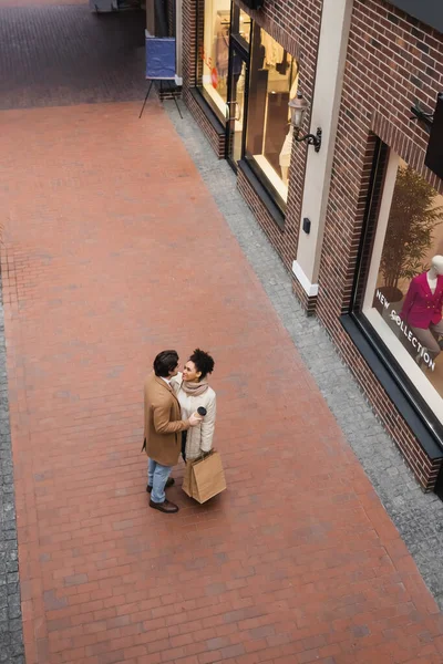 Hochwinkel-Ansicht des Mannes mit Pappbecher in der Nähe erfreut afrikanisch-amerikanische Freundin mit Einkäufen in Einkaufszentrum — Stockfoto