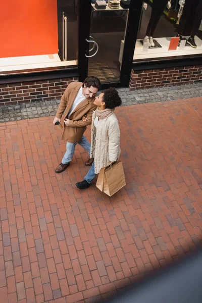 Vista de ángulo alto de alegre pareja multiétnica con compras caminando en el centro comercial - foto de stock