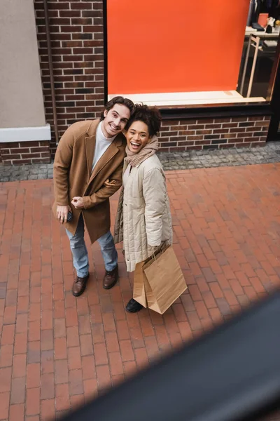 High angle view of joyful multiethnic couple with purchases looking at camera in mall — Stock Photo